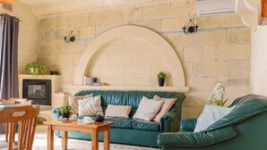 A cozy living room with a stone fireplace, a green sofa, a wooden table, and a comfortable chair. - Ta Dora 3 Bedroom Villa With Private Pool (Xaghra)