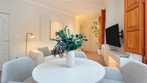 A modern living room with a round white table, grey sofas, a white vase with green plants, a floor lamp, and a flat-screen TV mounted on the wall. - Genteel Home Bib rambla IV (Granada)