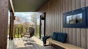 Een houten terras met een bank en rieten stoelen, een ingelijst beeld op de muur, en uitzicht op de omgeving.