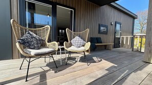 A wooden deck with wicker chairs and a bench, a sliding glass door, and a framed picture on the wall. - Chalet Near Sint Maartenszee Beach (Sint Maartensvlotbrug)