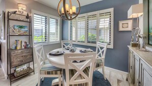 Un coin repas avec une table ronde, des chaises blanches et un lustre. On y trouve des fenêtres avec des volets et une bibliothèque agrémentée d’objets décoratifs.