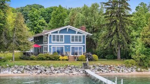 A house with a dock extending into a lake, surrounded by lush greenery and trees. - LakeHaus Vibes + Hot Tub (Traverse City)