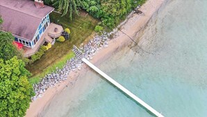 An aerial view of a beach with a long white pier extending into the water, surrounded by lush greenery and a house with a red roof. - LakeHaus Vibes + Hot Tub (Traverse City)