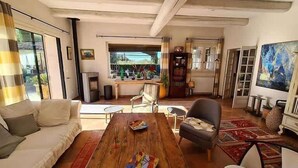 A spacious living room with a wooden coffee table, a sofa, and a chair. There is a fireplace, a bookshelf, and a large window with a view of the outdoors. - La Villa de Robion Su (Robion)