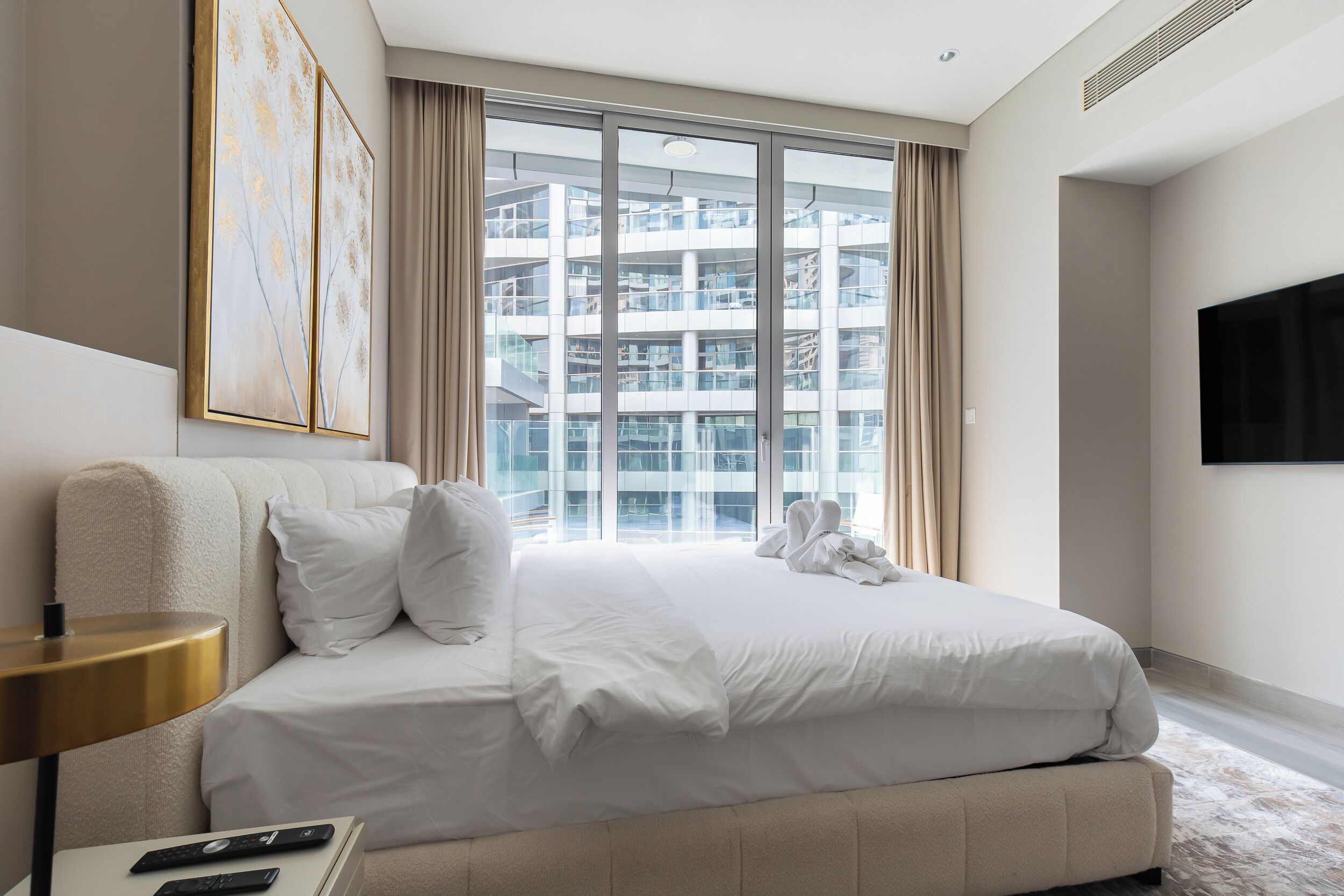 A modern hotel room with a large bed, a bedside table with a lamp, a flat-screen TV, and a view of a high-rise building through floor-to-ceiling windows.