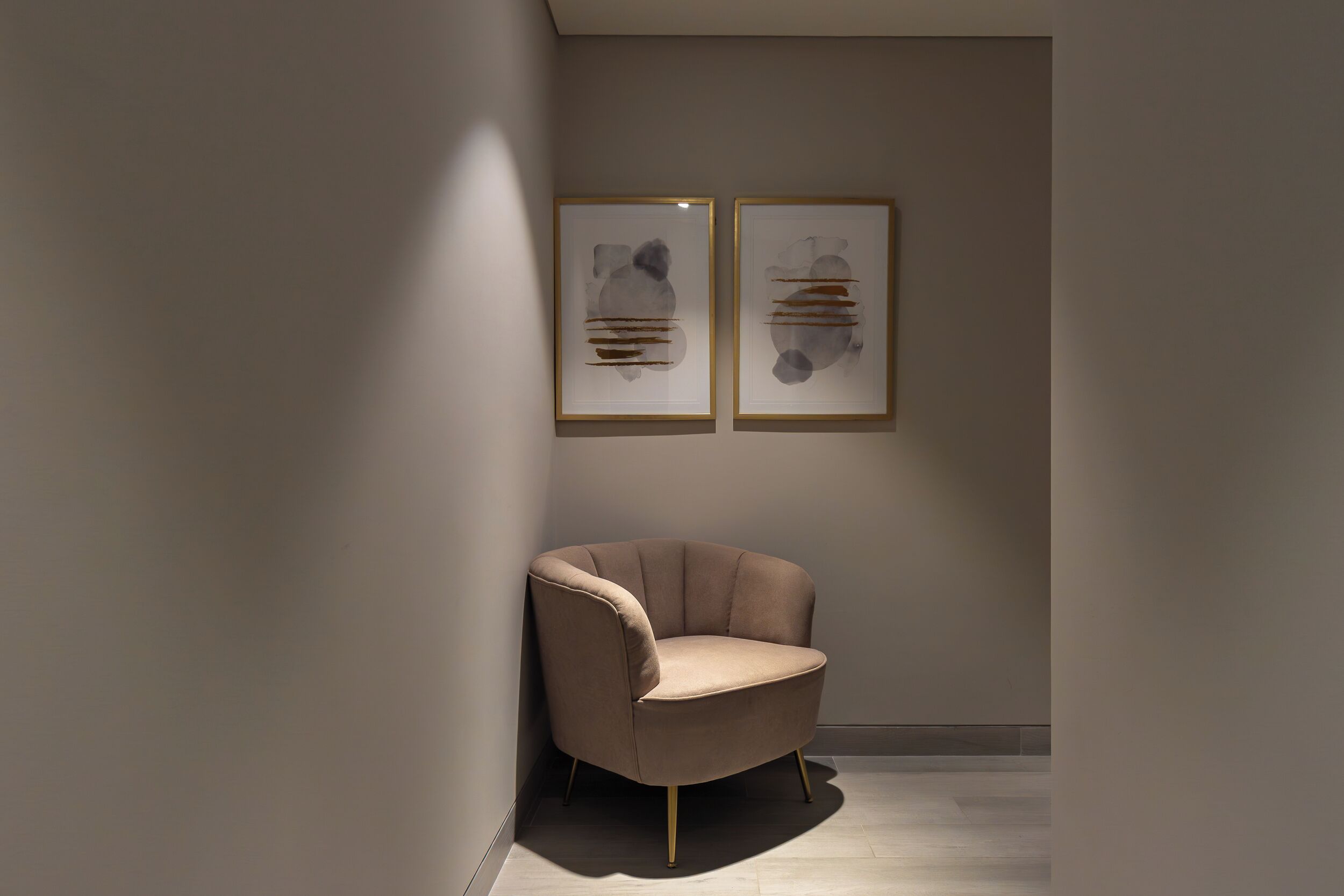 Una habitación moderna y minimalista con un sillón marrón, dos cuadros abstractos en la pared y un suelo de tonos claros.