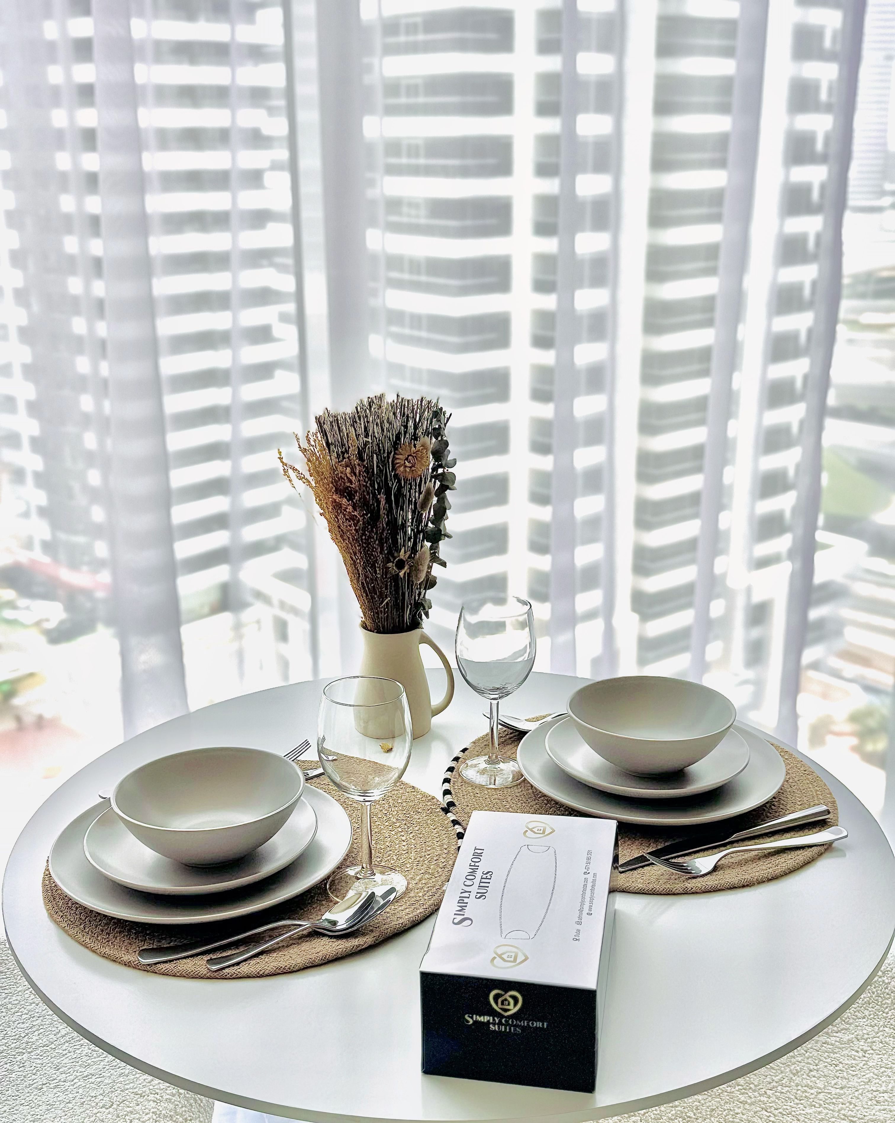A dining table set with white plates, bowls, and cutlery, a vase with dried flowers, and a menu on a placemat.
