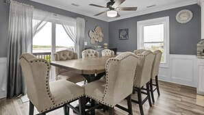 A dining room with a large table and six chairs, a ceiling fan, and a view through the window. - Waterfront Stay w/ Stunning Views, Beach Access, Outdoor Shower & Lift (North Topsail Beach)
