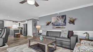 A modern living room with a grey sofa, a wooden coffee table, and a kitchen in the background. - Waterfront Stay w/ Stunning Views, Beach Access, Outdoor Shower & Lift (North Topsail Beach)