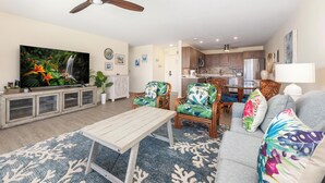 Un salon avec un téléviseur, une table basse en bois et des fauteuils à motifs tropicaux.