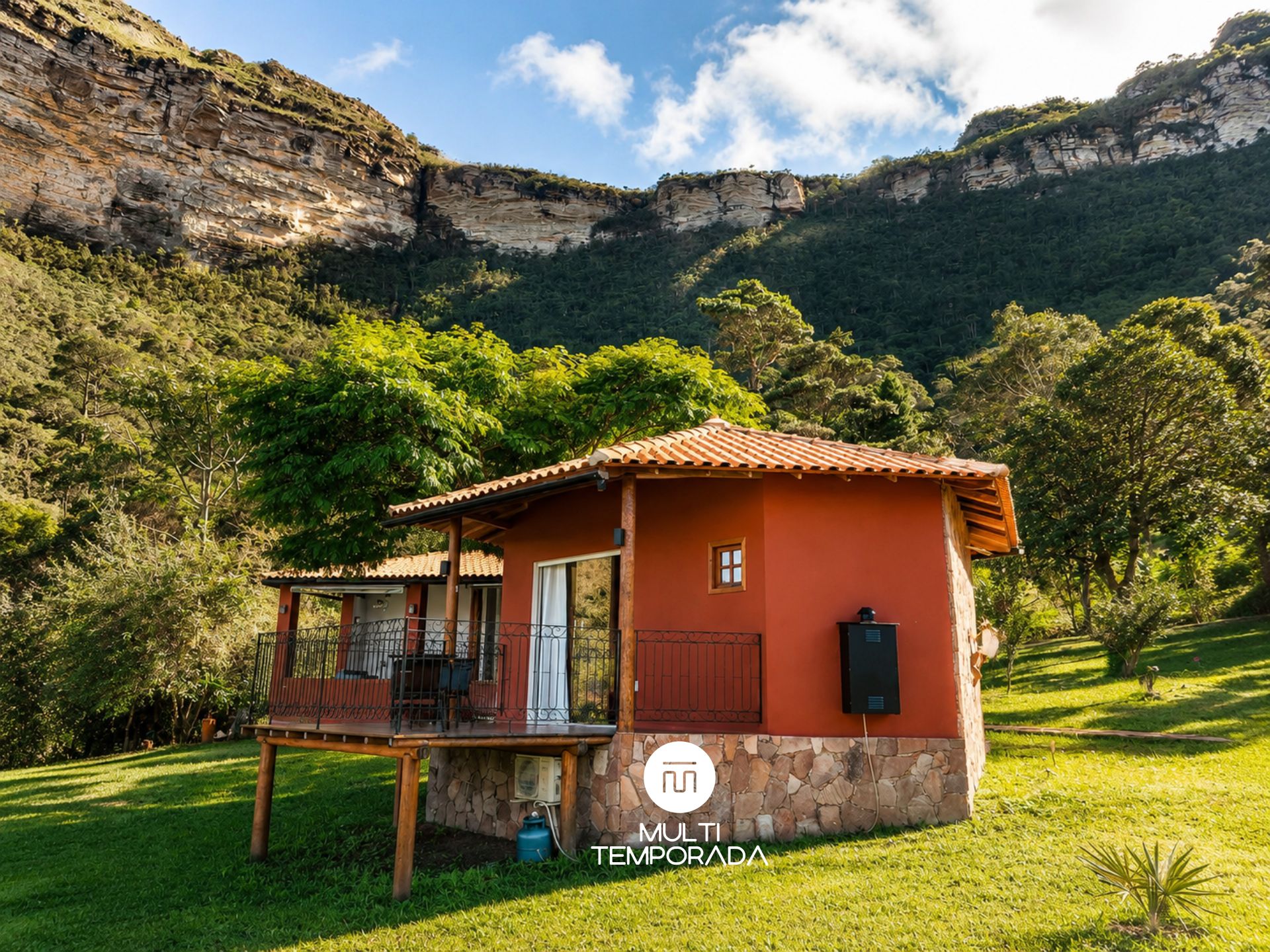 Um pequeno edifício térreo com telhado vermelho, varanda e alicerce de pedra, rodeado por vegetação exuberante e montanhas.