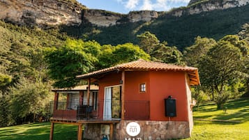 Um pequeno edifício térreo com telhado vermelho, varanda e alicerce de pedra, rodeado por vegetação exuberante e montanhas.