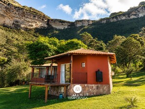 A small, single-story building with a red roof, a balcony, and a stone foundation, surrounded by lush greenery and mountains.