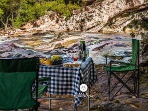 Mesa para comer al aire libre con mantel a cuadros, una botella y comida, con vista a un lecho rocoso de río.