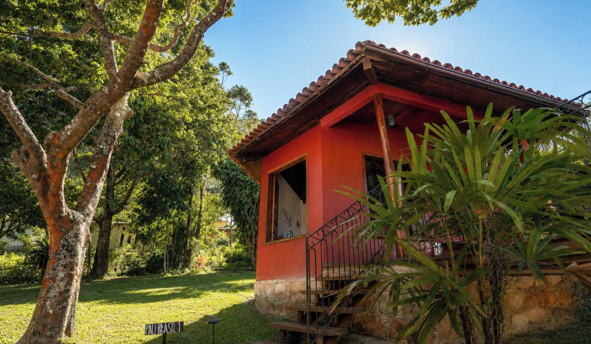 A small red building with a stone base, surrounded by lush greenery and a tree.