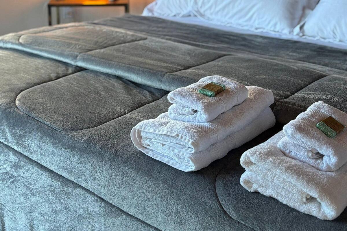 A neatly made bed with white pillows and a grey quilted headboard. A small table with a lit lamp is visible in the background.