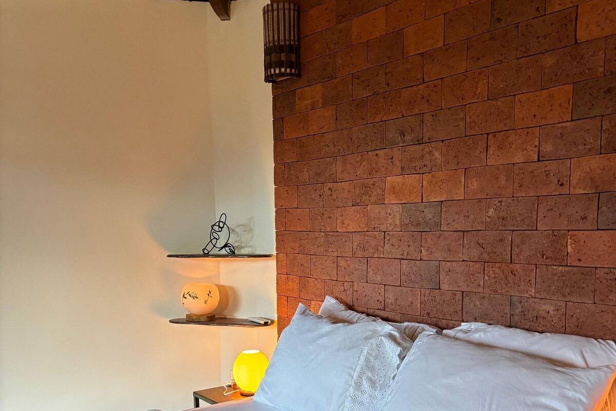 A neatly made bed with white linens and a beige blanket, a small bedside table with a lamp, and a brick wall with shelves above.
