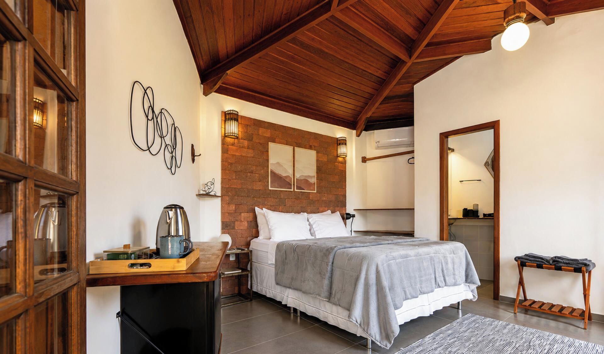 Quarto com cama, escrivaninha de madeira, cafeteira e parede de tijolos.