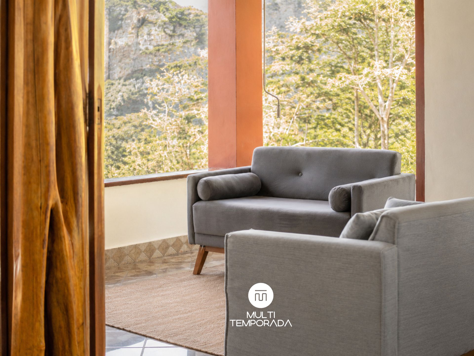 A modern living room with a grey sofa, a wooden floor, and a view of greenery through large windows.