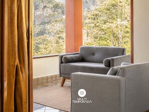 A modern living room with a grey sofa, a wooden floor, and a view of greenery through large windows.