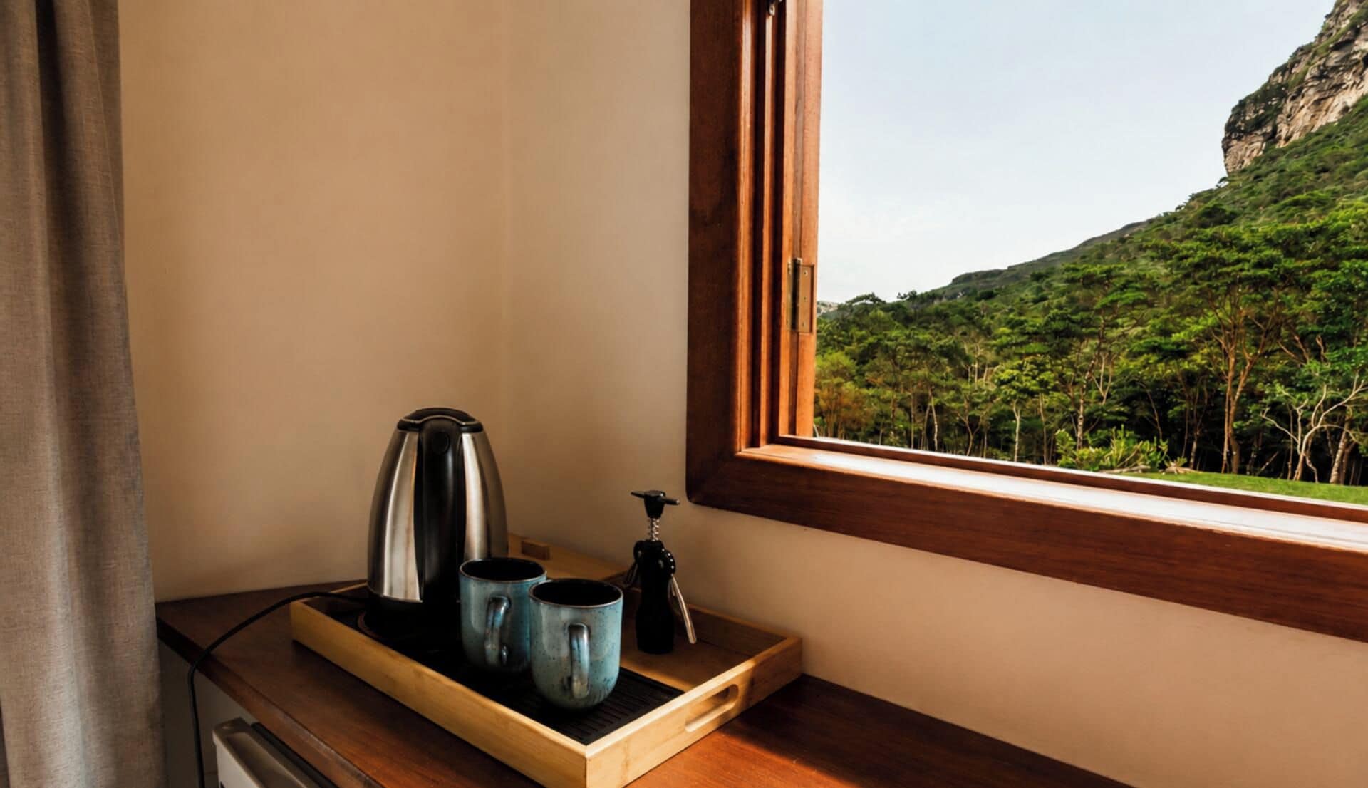 A wooden tray with a kettle and two mugs on a windowsill, overlooking a forested hillside.