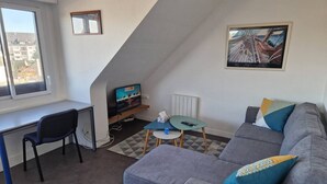 A small, modern living room with a grey sofa, a blue chair, a small table, and a TV. There is a window with a view of buildings outside. - Glaz - T1bis in the heart of Pllemeur (Ploemeur)