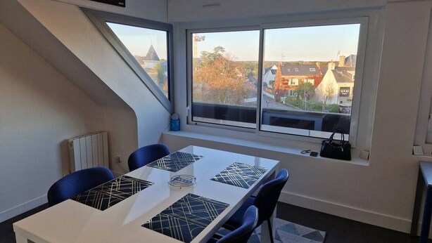 A modern room with a dining table, chairs, a window with a view of buildings, and a staircase. - Glaz - T1bis in the heart of Pllemeur (Ploemeur)