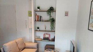 Ein modernes Wohnzimmer mit einem grauen Sofa, einem kleinen Hocker, einem runden Tisch mit einer Vase sowie einem Bücherregal mit Pflanzen und Büchern.
