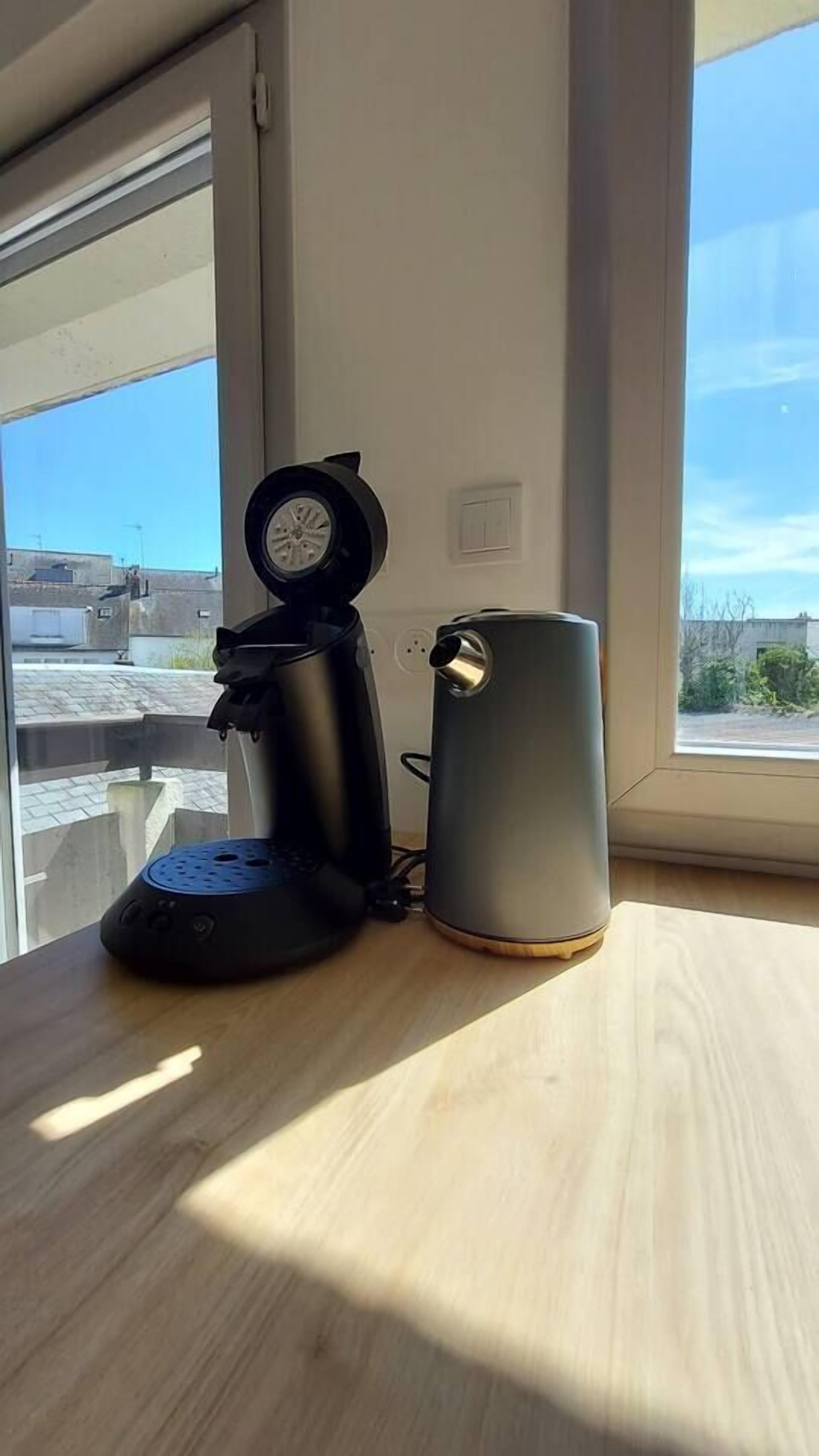 Une machine à café et une tasse sur une table en bois près d’une fenêtre donnant sur des immeubles.