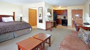 Habitación de hotel con cama, dos mesitas de noche de madera, una mesa de madera, un sofá, zona de cocina con armarios y electrodomésticos, y una puerta que da a otra habitación.