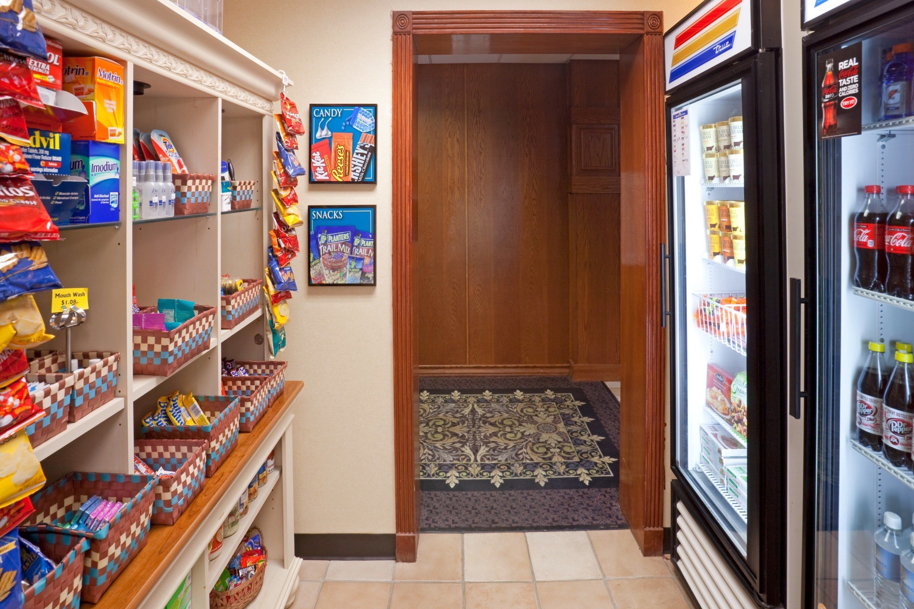 A convenience store with shelves stocked with snacks and candy, a refrigerator with beverages, and a wooden door leading to another room.
