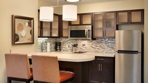A modern kitchen with dark wood cabinets, stainless steel appliances, and a white countertop. There are two chairs with patterned upholstery and a hanging light fixture above the counter.