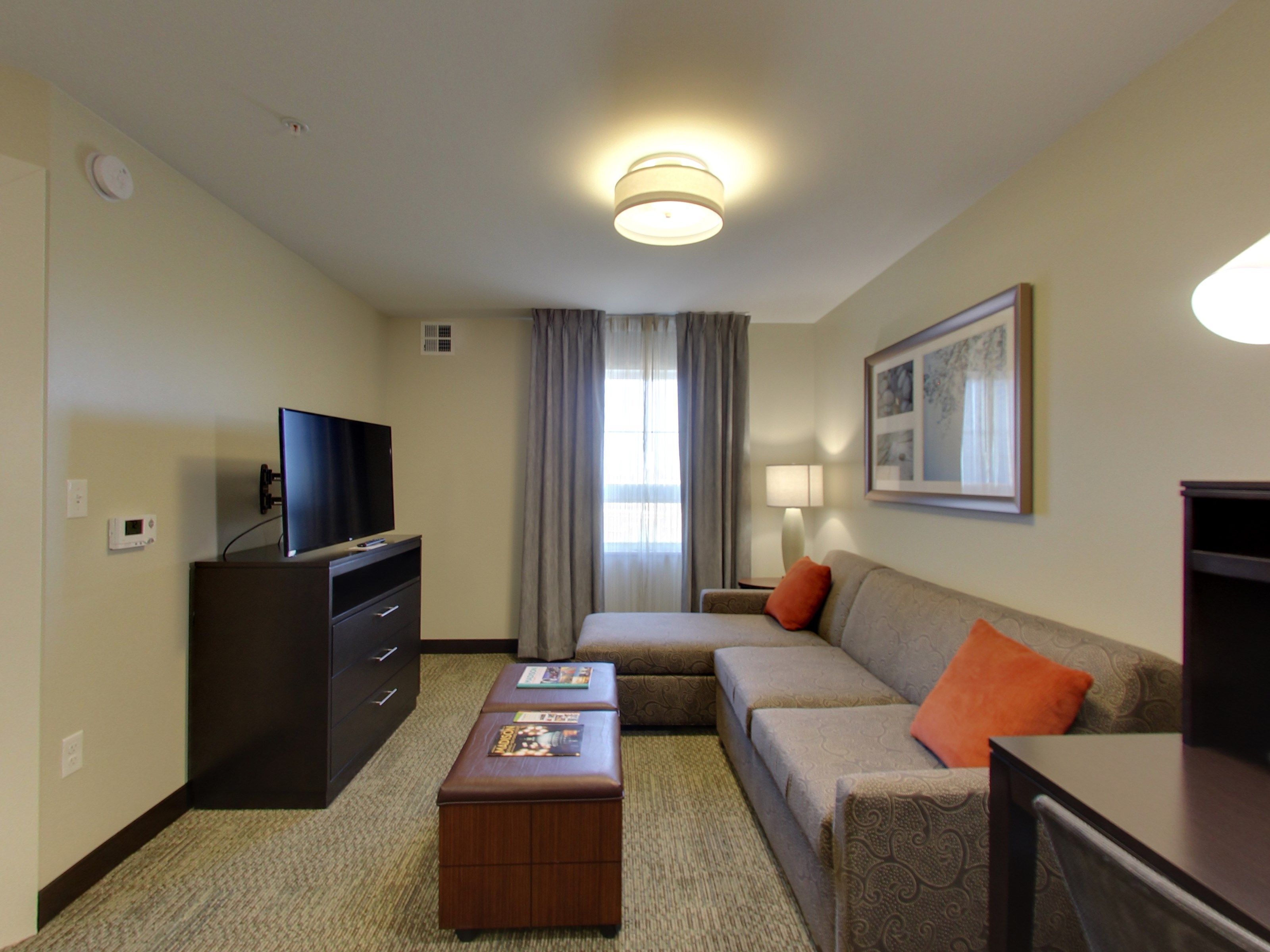 Een hotelkamer met een flatscreen televisie op een dressoir, een bank met een oranje kussen, een klein houten tafeltje met tijdschriften, een lamp en een ingelijst schilderij aan de muur.