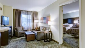 Een hotelkamer met een bank, fauteuil en een klein bureau met een lamp. Er is een televisie op een standaard en een bed met hoofdeinde.