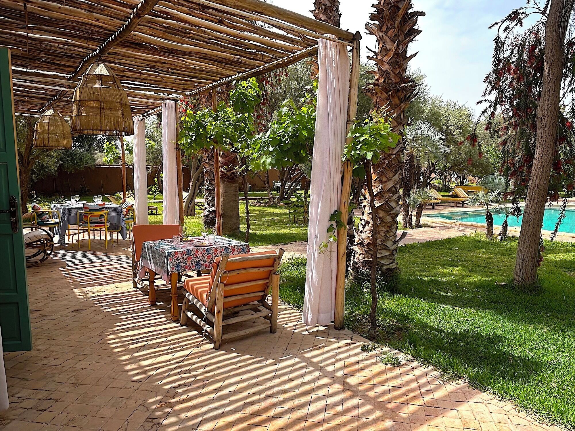 An outdoor patio with tables and chairs, a pergola with hanging lights, a pool, and surrounding greenery.