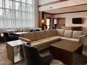 A hotel lobby with a sectional sofa, round tables, and a flat-screen TV mounted on the wall.