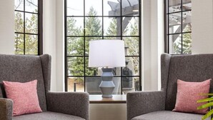 Two grey armchairs with pink cushions, a white table lamp, and a window with a view of trees. - Cozy Studio | Free Breakfast. Pool & Gym. (Maple Grove)