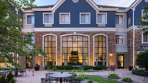 A two-story building with a stone facade, large arched windows, and a patio area with outdoor seating. - Cozy Studio | Free Breakfast. Pool & Gym. (Maple Grove)
