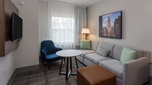 Une chambre d’hôtel avec un canapé, un fauteuil, une petite table, un téléviseur, une lampe et un tableau au mur.
