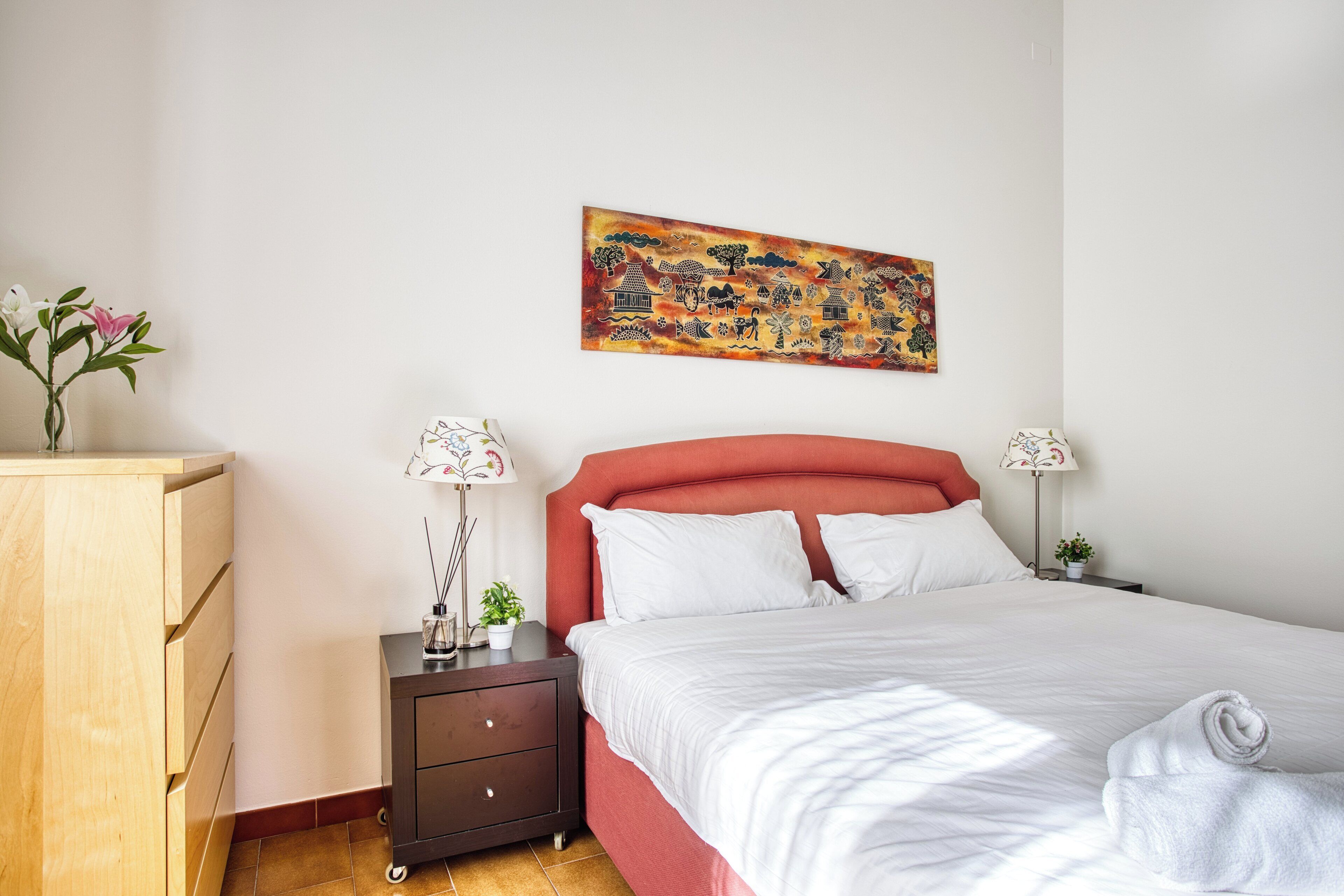 Un dormitorio con una cama, dos mesitas de noche, un tocador, un aparador de madera y un cuadro de colores en la pared.