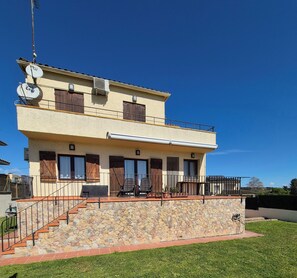 A two-story building with a stone staircase, a balcony with chairs, and a satellite dish.