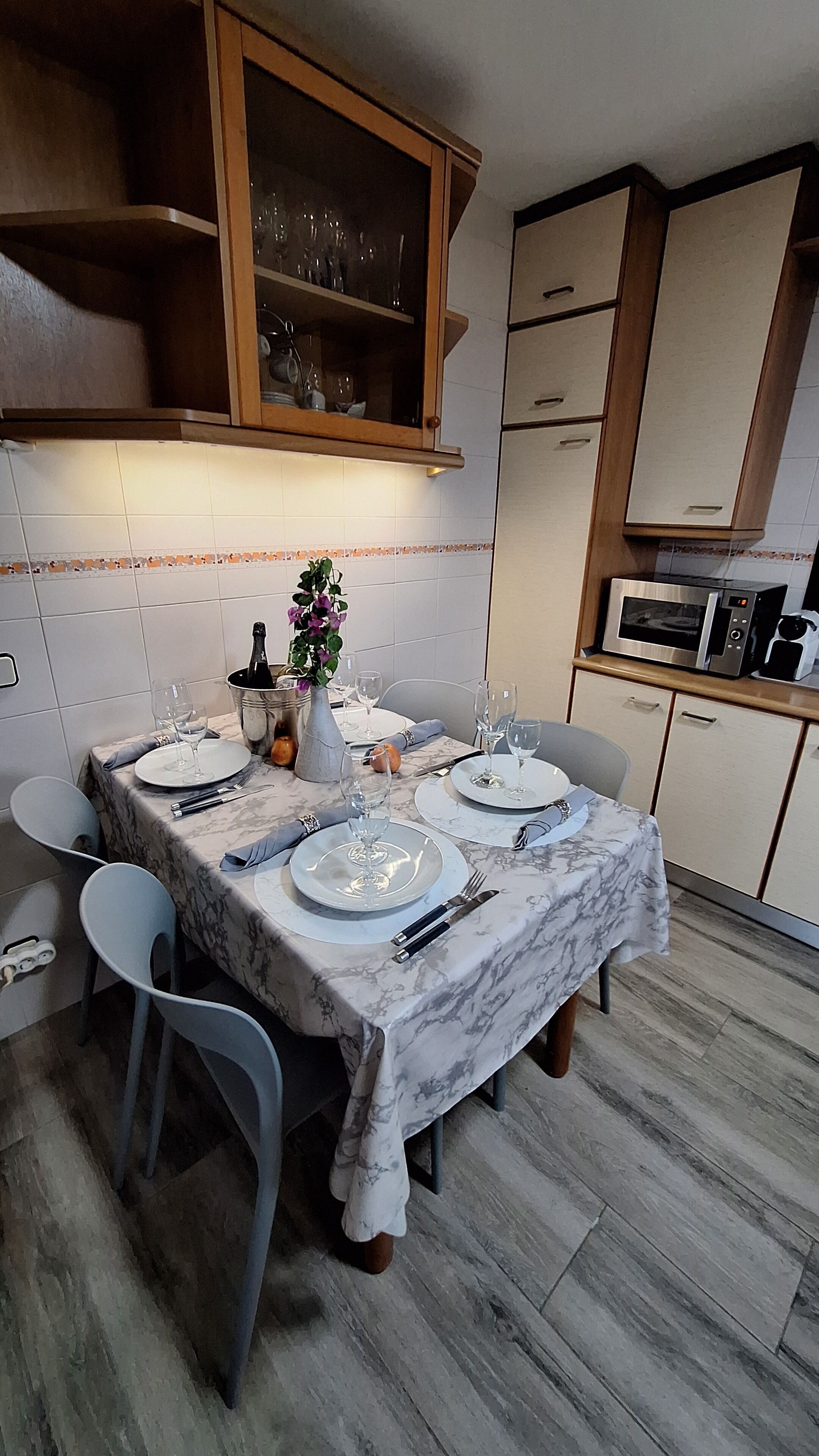 Un coin repas avec une table en marbre dressée pour quatre personnes, comprenant des assiettes, des verres et des couverts. On y trouve deux chaises, un vase avec des fleurs et une bouteille de champagne. La pièce est dotée de meubles en bois et un four à micro-ondes est visible au fond.