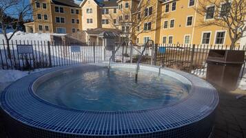 A hot tub in a courtyard with a snow-covered ground, surrounded by a fence and a building with multiple windows.