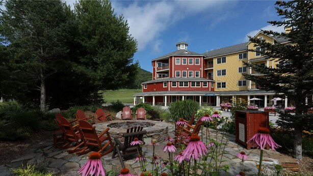 A multi-story building with a red and yellow facade, surrounded by lush greenery and flowering plants. - Okemo | Studio Condo + Close to All Amenities (Ludlow)