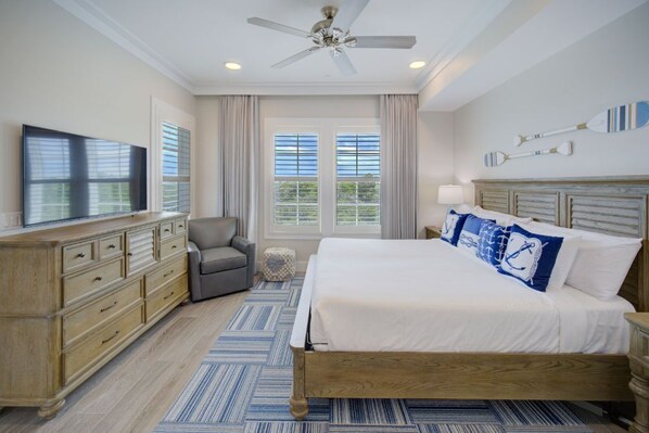 A bedroom with a large bed, a television, a ceiling fan, and a view of the outdoors through the windows.