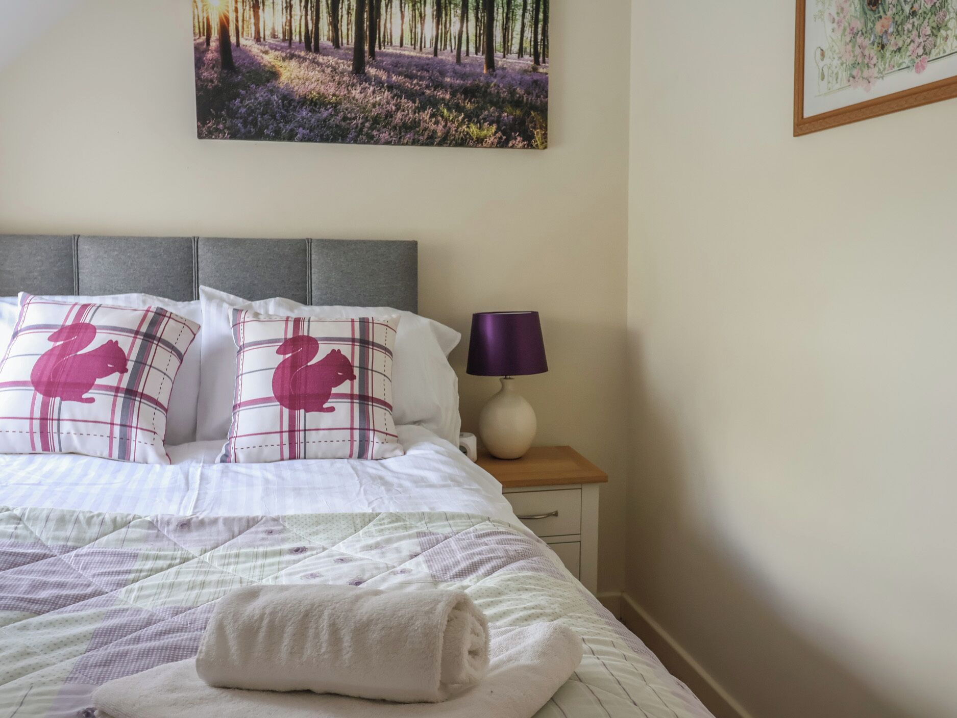 A neatly made bed with a plaid pillow, a rolled white towel, and a nightstand with a lamp.