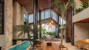 Amplia zona de piscina interior con mesa y sillas de madera, rodeada de exuberante vegetación y un muro de piedra.