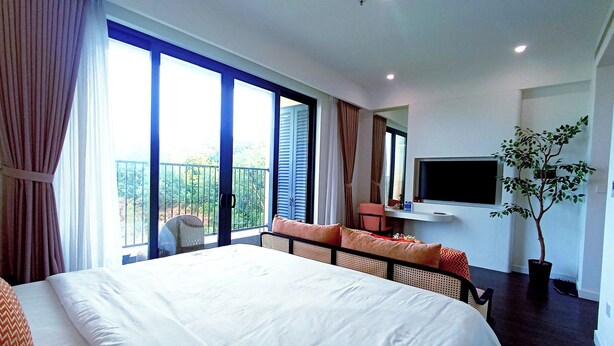 A hotel room with a large bed, a TV mounted on the wall, a balcony with curtains, and a potted plant.