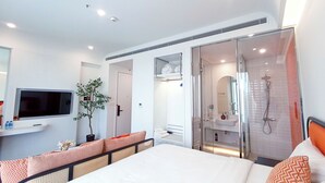 A modern hotel room with a large bed, a TV, a plant, and a bathroom with a shower and sink.