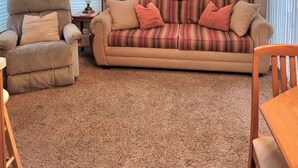 Una sala de estar con un sofá color beige, un sillón de madera y una mesa de comedor de madera. Hay un ventilador de techo, una ventana con cortinas y una lámpara sobre una mesita.